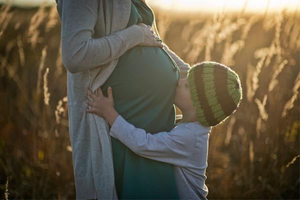 Una mujer embarazada acompañada por su hijo pequeño, que abraza su vientre, en un campo iluminado por el sol