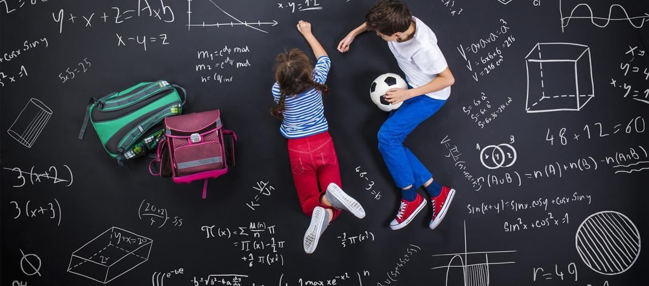 Dos niños acostados sobre un tablero cubierto de fórmulas matemáticas, con morrales y un balón de fútbol